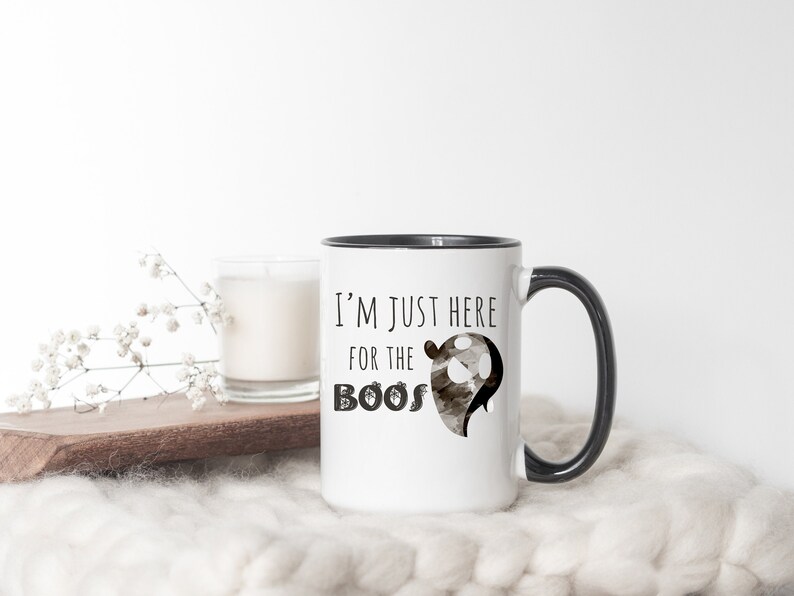 May include: White ceramic mug with a black rim and handle. The mug has a black and white graphic of a ghost with the text "I'm just here for the boos".