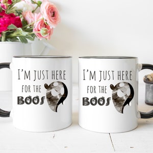 May include: Two white ceramic mugs with black handles and black rims. The mugs have a black and white ghost graphic and the text "I'm just here for the boos".