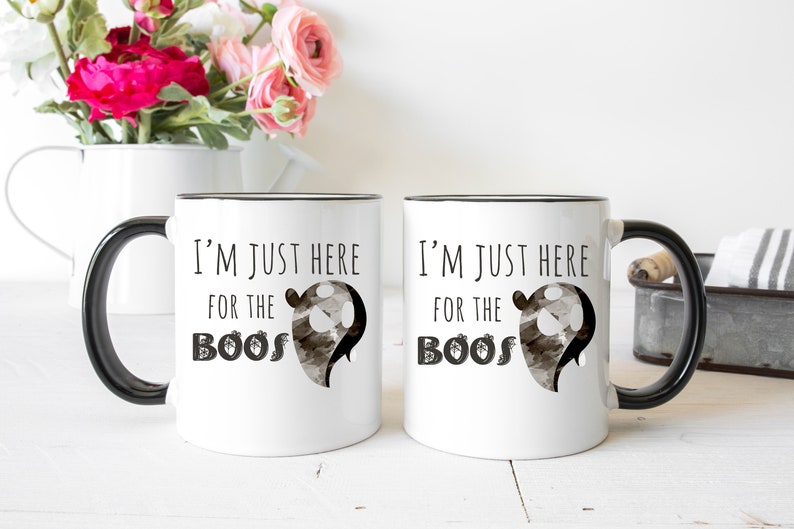 May include: Two white ceramic mugs with black handles and black rims. The mugs have a black and white ghost graphic and the text "I'm just here for the boos".