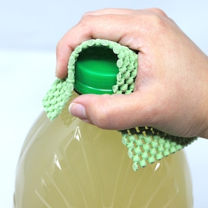 May include: A clear plastic bottle with a green lid is being held by a hand wearing a green dishcloth. The bottle is filled with a light yellow liquid.