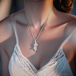 May include: A gold-toned necklace featuring a slender, elongated pendant with an iridescent, light-reflecting centre stone. The pendant is accented by a delicate, spiralling gold-toned metal detail encrusted with small, clear, sparkling stones.