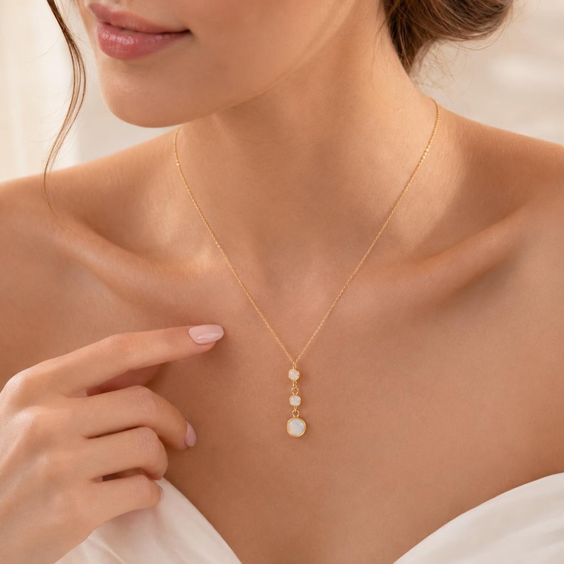 May include: A delicate gold chain necklace featuring a vertical drop pendant with three round, iridescent white stones set in gold bezels. The stones are arranged in a descending size order, suspended from a thin, polished gold link chain.