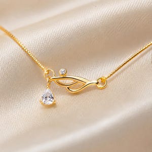 May include: A gold-toned necklace featuring a delicate box chain and a central pendant. The pendant consists of a curved, abstract metal element adorned with a small round clear stone and a dangling pear-shaped clear crystal drop charm.
