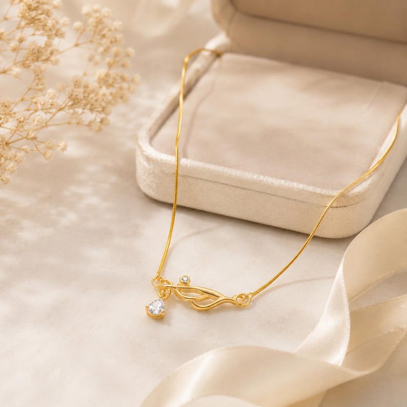 May include: A gold-toned necklace featuring a delicate chain and a central pendant shaped like a stylised stethoscope. The pendant is accented with two small, clear, round-cut stones, one set within the metal frame and one dangling teardrop-shaped stone.