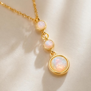 May include: A gold chain necklace featuring a vertical drop design with three round, bezel-set opals. The stones display iridescent flashes of color, and the metal has a polished gold finish. The pendant consists of three circular links connected in a row.