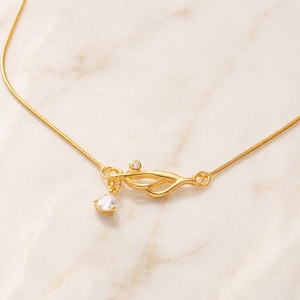 May include: A gold-toned necklace featuring a delicate snake chain and a central abstract metal element. This centrepiece is accented with a small, round, clear stone and a dangling, pear-shaped, clear faceted stone that catches the light.