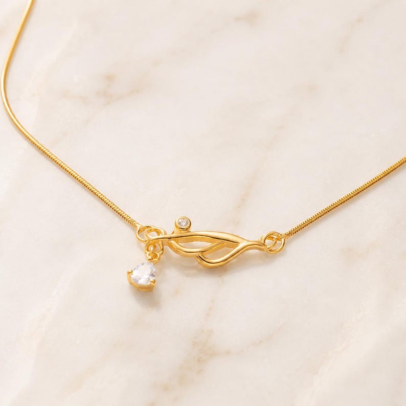 May include: A gold-toned necklace featuring a delicate snake chain and a central abstract metal element. This centrepiece is accented with a small, round, clear stone and a dangling, pear-shaped, clear faceted stone that catches the light.