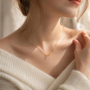 May include: A delicate gold chain necklace featuring a small, intricate leaf-shaped pendant with a single clear, sparkling stone suspended from the centre. The thin, polished gold chain rests against the collarbone, highlighting the metallic finish.