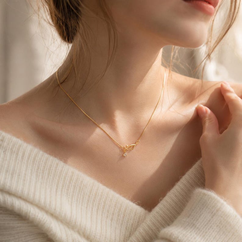 May include: A delicate gold chain necklace featuring a small, intricate leaf-shaped pendant with a single clear, sparkling stone suspended from the centre. The thin, polished gold chain rests against the collarbone, highlighting the metallic finish.
