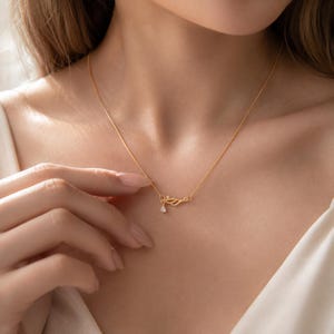 May include: A delicate gold chain necklace featuring a small, stylised wing-shaped pendant. The pendant is accented with a single, tiny, clear teardrop-shaped gemstone hanging from the lower edge of the wing design, creating a subtle and refined look.
