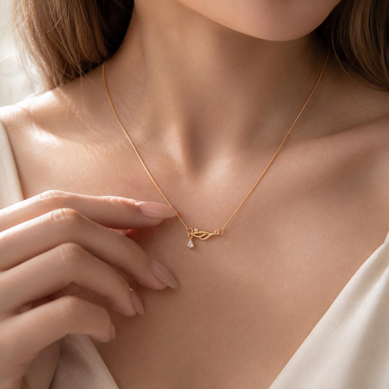 May include: A delicate gold chain necklace featuring a small, stylised wing-shaped pendant. The pendant is accented with a single, tiny, clear teardrop-shaped gemstone hanging from the lower edge of the wing design, creating a subtle and refined look.