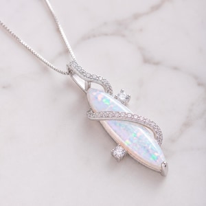 Könnte beinhalten: Eine silberfarbene Anhängerkette mit einem langen, marquiseförmigen weißen Opal mit irisierenden Farbblitzen. Der Opal wird von einem geschwungenen Metallrahmen akzentuiert, der mit kleinen klaren Steinen und zwei größeren runden Steinen besetzt ist.