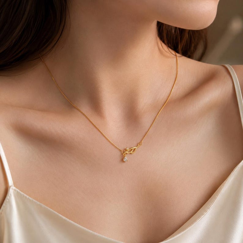 May include: A delicate gold chain necklace featuring a small, asymmetrical leaf-shaped pendant. The pendant is accented with a single, tiny clear gemstone set within the metal frame and a small round clear stone dangling from the bottom.