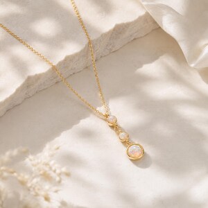 May include: A gold-tone chain necklace featuring a vertical drop pendant with three round, bezel-set iridescent opal-like stones. The stones are arranged in a descending size order, with the largest at the bottom, all connected by small gold rings.