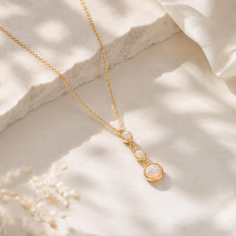 May include: A gold-tone chain necklace featuring a vertical drop pendant with three round, bezel-set iridescent opal-like stones. The stones are arranged in a descending size order, with the largest at the bottom, all connected by small gold rings.