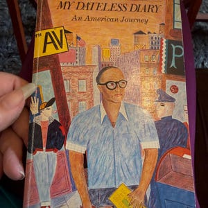 May include: A paperback book titled My Dateless Diary: An American Journey by R.K. Narayan. The cover features a colorful illustration of a person wearing glasses and a blue shirt, standing in a city scene with buildings, a street sign, and people.