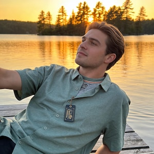 May include: A light green button-down shirt with a tag attached. The shirt is worn over a gray t-shirt. The tag has the word "Poutin" on it. The background shows a lake and a sunset.