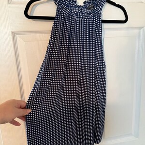 Pu&ograve; includere: Un top senza maniche con motivo a quadretti blu navy e bianco. Il top ha uno scollo a balze ed &egrave; appeso a una gruccia nera. Il tessuto &egrave; leggero e sembra essere un materiale morbido.