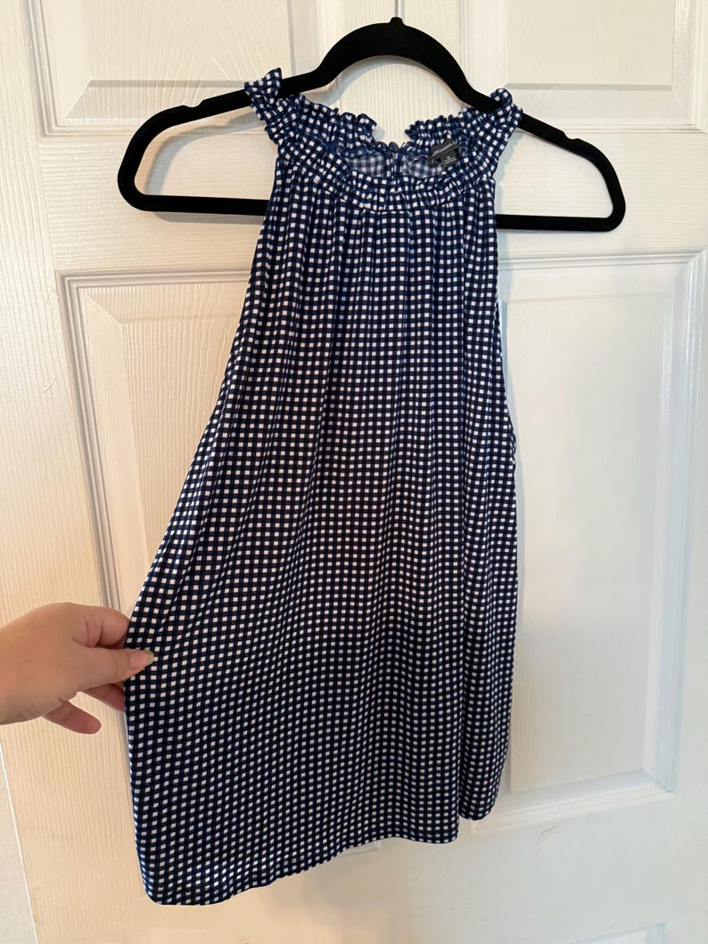 Pu&ograve; includere: Un top senza maniche con motivo a quadretti blu navy e bianco. Il top ha uno scollo a balze ed &egrave; appeso a una gruccia nera. Il tessuto &egrave; leggero e sembra essere un materiale morbido.