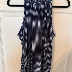Pu&ograve; includere: Un top senza maniche con stampa a quadretti blu navy e bianca con scollatura arricciata. Il top &egrave; appeso a una gruccia nera contro una porta bianca.