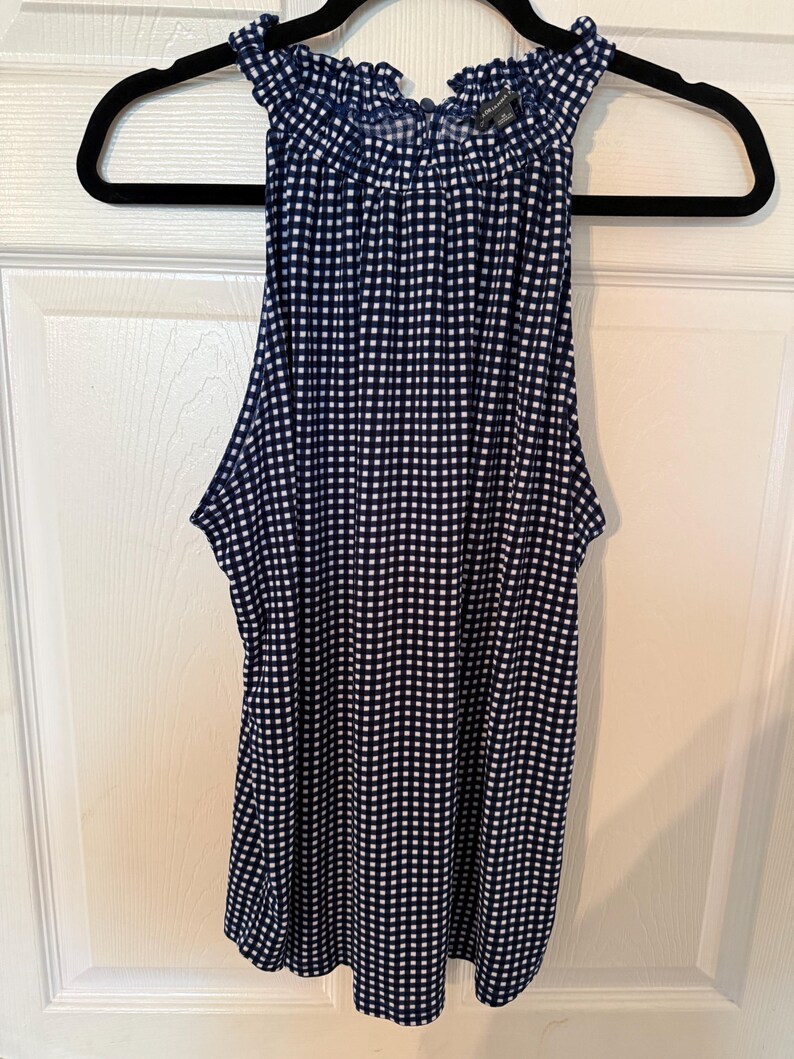 Pu&ograve; includere: Un top senza maniche con stampa a quadretti blu navy e bianca con scollatura arricciata. Il top &egrave; appeso a una gruccia nera contro una porta bianca.