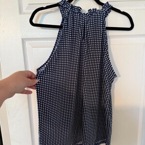 Pu&ograve; includere: Top senza maniche con stampa a quadretti blu navy e bianca con scollatura arricciata. Il top &egrave; appeso a una gruccia nera contro una porta bianca. Il top &egrave; realizzato in tessuto leggero.