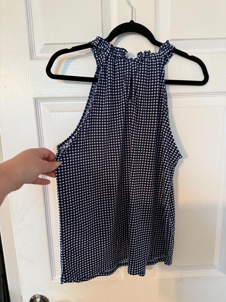 Pu&ograve; includere: Top senza maniche con stampa a quadretti blu navy e bianca con scollatura arricciata. Il top &egrave; appeso a una gruccia nera contro una porta bianca. Il top &egrave; realizzato in tessuto leggero.
