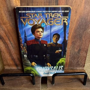 May include: A Star Trek Voyager book titled "Chrysalis" by David Niall Wilson. The cover features a blue background with images of two people in Star Trek uniforms. The book is held in a wooden book stand.