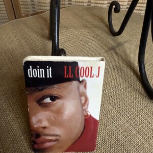 May include: A small, rectangular item featuring a close-up image of LL Cool J. The text "doin it" and "LL COOL J" are printed on the top. The item is set against a neutral background, with a black metal decorative element in the background.
