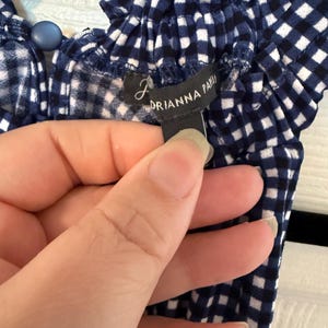 Pu&ograve; includere: Primo piano di un top a quadretti blu e bianchi. Il top ha uno scollo a balze e un bottone blu. L'etichetta recita "DRIANNA PAPI".
