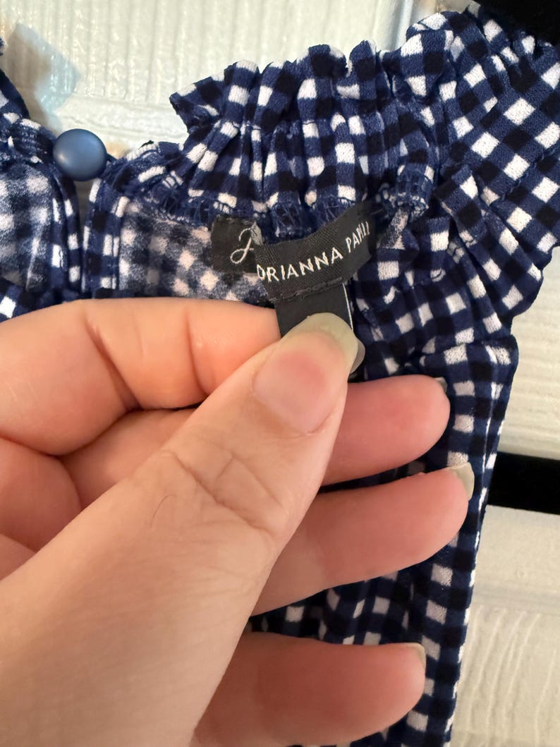 Pu&ograve; includere: Primo piano di un top a quadretti blu e bianchi. Il top ha uno scollo a balze e un bottone blu. L'etichetta recita "DRIANNA PAPI".