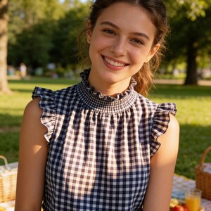 Pu&ograve; includere: Un top senza maniche con un motivo a quadretti vichy blu navy e bianco. Il top ha uno scollo e giromanica a balze. La persona sorride in un ambiente di parco con alberi e un cestino da picnic sullo sfondo.