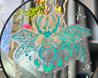 Puede incluir: Un espejo redondo con marco negro, que presenta una pegatina holográfica de un escarabajo verde con alas y lunas. La pegatina tiene un acabado iridiscente.