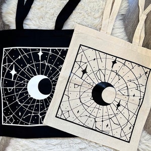 Op de afbeelding: Twee tote bags met zwarte en witte hemelontwerpen. Eén tas is zwart met witte ontwerpen, en de andere is beige met zwarte ontwerpen. Beide tassen hebben een halve maan, sterren en een sterrenbeeldpatroon.