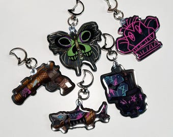 Puede incluir: Cinco llaveros coloridos con diferentes diseños. Una mariposa verde y negra, una calavera rosa y negra, una pistola negra y blanca, un tiburón negro y blanco y un robot negro y blanco.