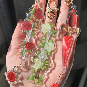 Peut inclure: Trois porte-clés en forme d'épée en acrylique avec des accessoires dorés. L'un des porte-clés présente des roses rouges, un autre des fleurs blanches et le troisième un motif de lame rouge. Les porte-clés sont tenus dans une main.