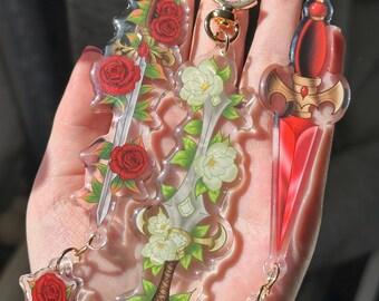 Puede incluir: Tres llaveros acrílicos con forma de espada con herrajes dorados. Un llavero presenta rosas rojas, otro flores blancas y el tercero un diseño de hoja roja. Los llaveros se sostienen en una mano.