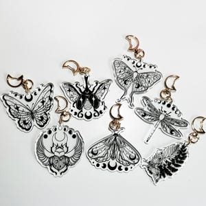 Op de afbeelding: Zeven transparante acryl sleutelhangers met zwart-wit illustraties van insecten en halve manen. De insecten omvatten een vlinder, een libel, een nachtvlinder en een kever.