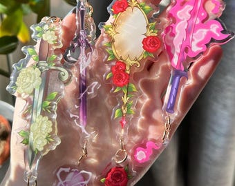 Puede incluir: Una colección de llaveros de acrílico transparente con diseños de espadas y espejos. Los llaveros tienen herrajes dorados y están decorados con motivos florales y de llamas en tonos rosa, morado, verde y rojo.