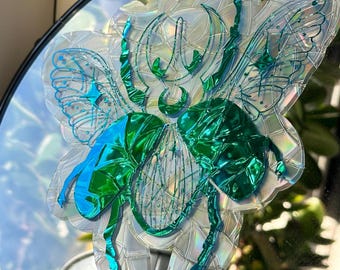 Puede incluir: Espejo decorativo iridiscente con un diseño de insecto grande y estilizado. El insecto presenta alas y segmentos corporales en tonos verdes y azules, con una superficie reflectante y texturizada. El espejo es redondo con un marco negro.