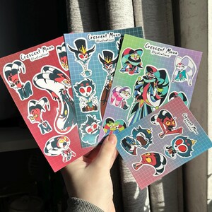 Op de afbeelding: Vier vellen met stickers met kleurrijke cartoonfiguren uit de animatieserie "Helluva Boss". De stickers tonen personages als Blitzo, Stolas en Moxxie. Elk vel heeft bovenaan de tekst "Crescent Moon Illustrations".