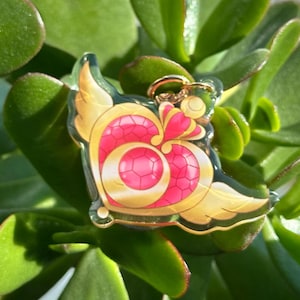 Op de afbeelding: Een gouden en roze hartvormige hanger met vleugels, een kroon en een cirkel in het midden. De hanger is gemaakt van helder acryl en heeft een gouden metalen lus aan de bovenkant.