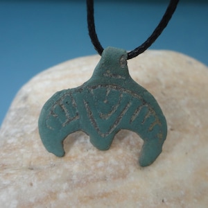 Puede incluir: Collar con colgante de color turquesa con cordón negro. El colgante tiene una forma curva estilizada con patrones geométricos grabados. El collar se muestra sobre una piedra clara y texturizada sobre un fondo azul.