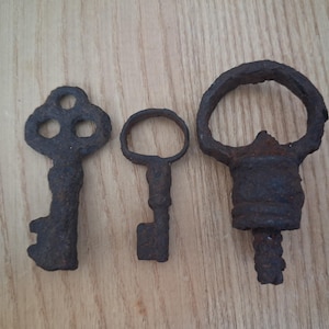 Puede incluir: Tres llaves antiguas de hierro de diferentes tamaños, dispuestas sobre una superficie de madera clara. Las llaves tienen un aspecto oscuro y envejecido, con óxido visible y diseños intrincados. La llave de la izquierda tiene tres agujeros circulares.