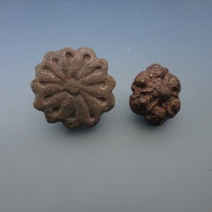 May include: Two antique bronze buttons with a floral design. The larger button has a more intricate design with a central cross shape. The smaller button has a more simple design with a raised center.