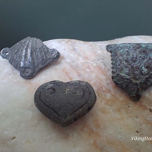 May include: Three weathered metal artifacts including a grey shell-shaped object with side loops, a dark grey heart-shaped piece with an engraved design, and a rectangular green-patinated metal fragment with an embossed pattern. Text: VikingHouseFind.