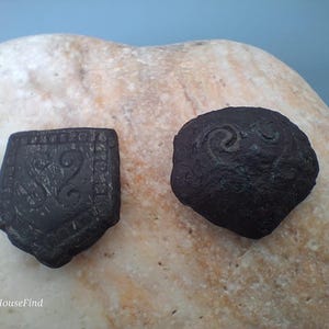 Könnte beinhalten: Zwei dunkle, möglicherweise antike Objekte liegen auf einem hellen Stein. Eines ist schildförmig mit einer Zahl 2 und anderen Designs. Das andere ist rund mit einem Wirbelmuster. Die Objekte sind dunkelbraun oder schwarz.