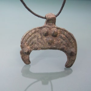 May include: A brown metal pendant with a crescent moon shape and three raised dots. The pendant is attached to a black cord.