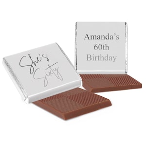 May include: Two square milk chocolate bars wrapped in silver foil. One wrapper features the text "She's Sixty" in a cursive script, while the other displays the text "Amanda's 60th Birthday" in a clean, sans-serif font. The chocolate surface is textured.