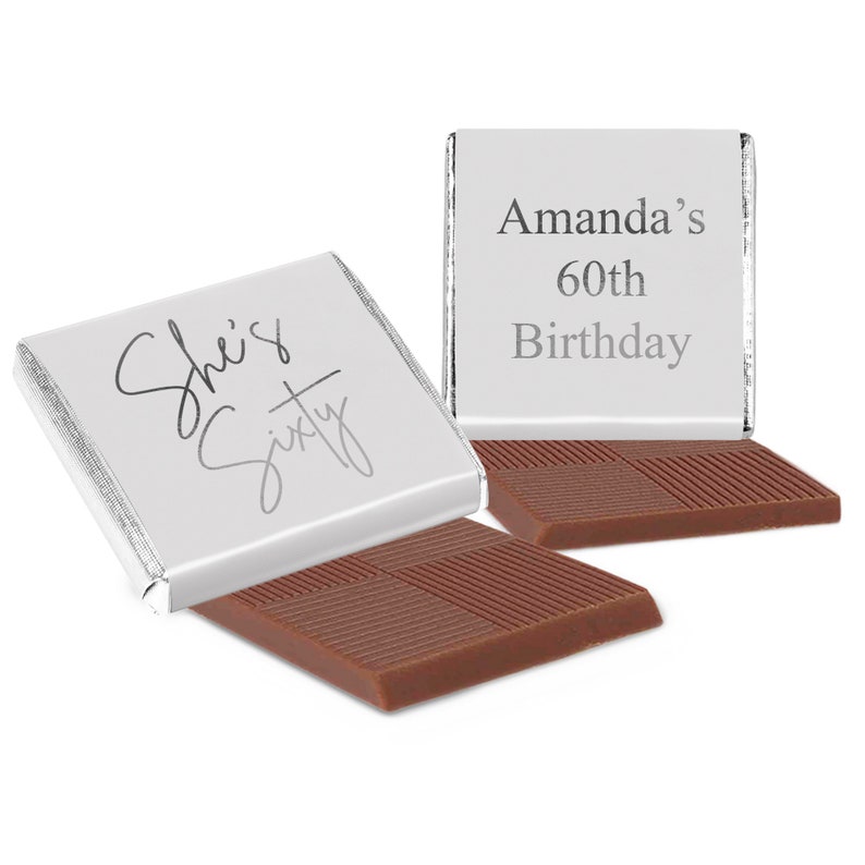 May include: Two square milk chocolate bars wrapped in silver foil. One wrapper features the text "She's Sixty" in a cursive script, while the other displays the text "Amanda's 60th Birthday" in a clean, sans-serif font. The chocolate surface is textured.